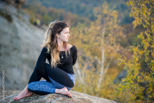 Attractive female is practicing yoga and doing asana Arha Matsyendrasana on the top of the high rocky mountain. Autumn forests, rocks and hills on the background photo