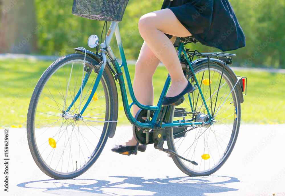 Fototapeta premium Girl on bicycle wearing on black short dress. Young Woman riding along road on green spring outdoor Park. Sporty young girl riding a bicycle on a sunny morning