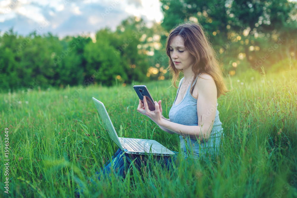 Woman sitting on a green meadow on the background of sunset. Working on laptop wireless and looking at the phone.