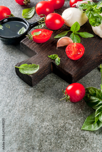 Farm raw organic products. Preparation of dinner in the Italian style. Ingredients for caprese salad, pasta, pizza. Basil, tomatoes, mozzarella cheese, olive oil on dark grey stone table. Copy space