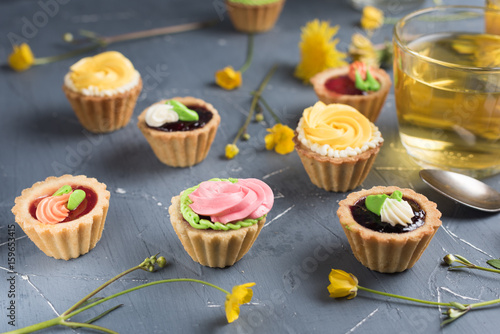 Colorful cupcakes on grey plate and table. Cakes with cream and jam. Cup of green tea and yellow flowers on a background