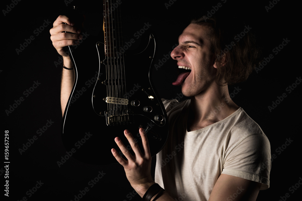 Music and creativity. Handsome young man in a T-shirt, with an electric guitar, sticks out his tongue, on a black isolated background. Horizontal frame