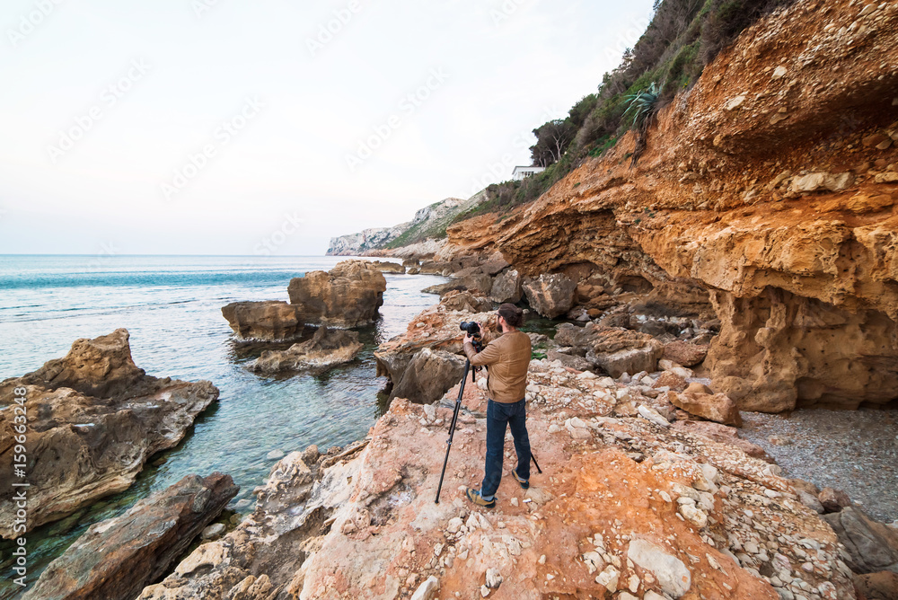 Young male photographer standing on rocks with tripod and making photos of sea Young male photographer standing on rocks with tripod and making photos of sea