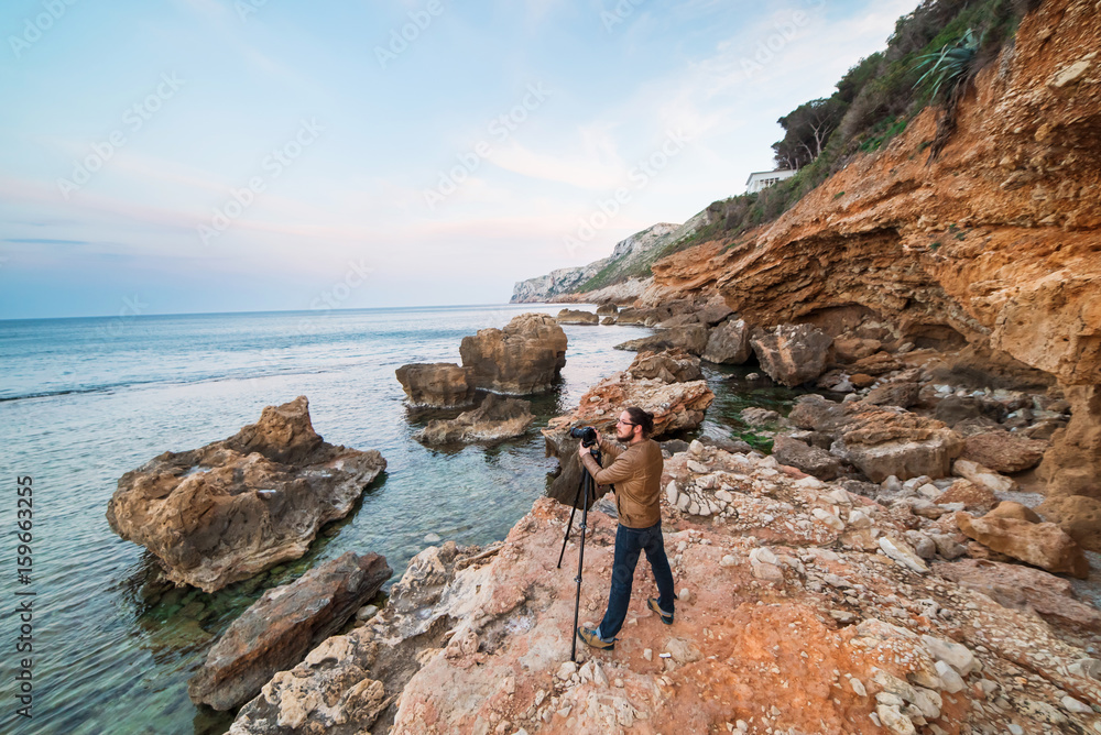 Young male photographer standing on rocks with tripod and making photos of sea