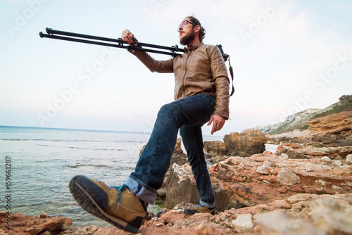 Young male photographer walking on rock beach with camera on tripod