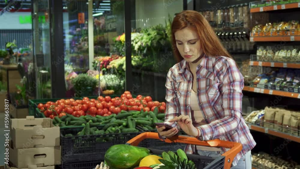 Attractive young couple checking their purchases according to the shopping list on the smartphone. Handsome blond man embracing his girlfriend at the supermarket. Pretty redhead girl and her boyfriend Attractive young couple checking their purchases according to the shopping list on the smartphone. Handsome blond man embracing his girlfriend at the supermarket. Pretty redhead girl and her boyfriend