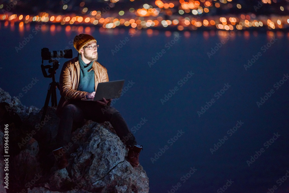 Young dreamy photographer sitting on precipice and working on laptop with view of sea and mountains