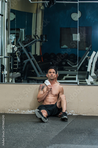 Middle age muscular man resting on gym floor with towel draped across shoulder.