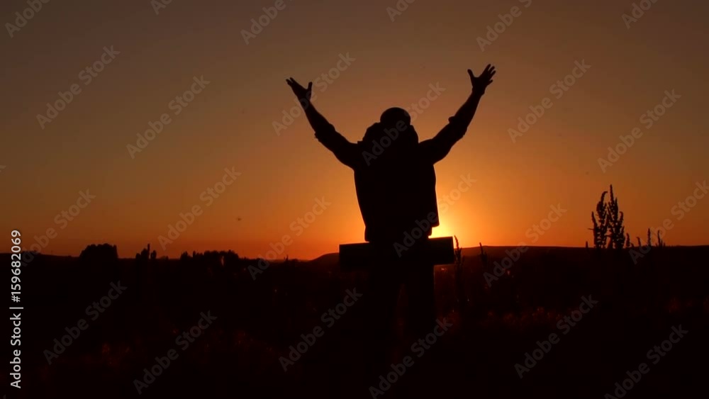 Silhouette of a traveler with a large backpack lifts hands to the sky standing on top of a mountain at sunset, slow motion. Silhouette of a traveler with a large backpack lifts hands to the sky standing on top of a mountain at sunset, slow motion.