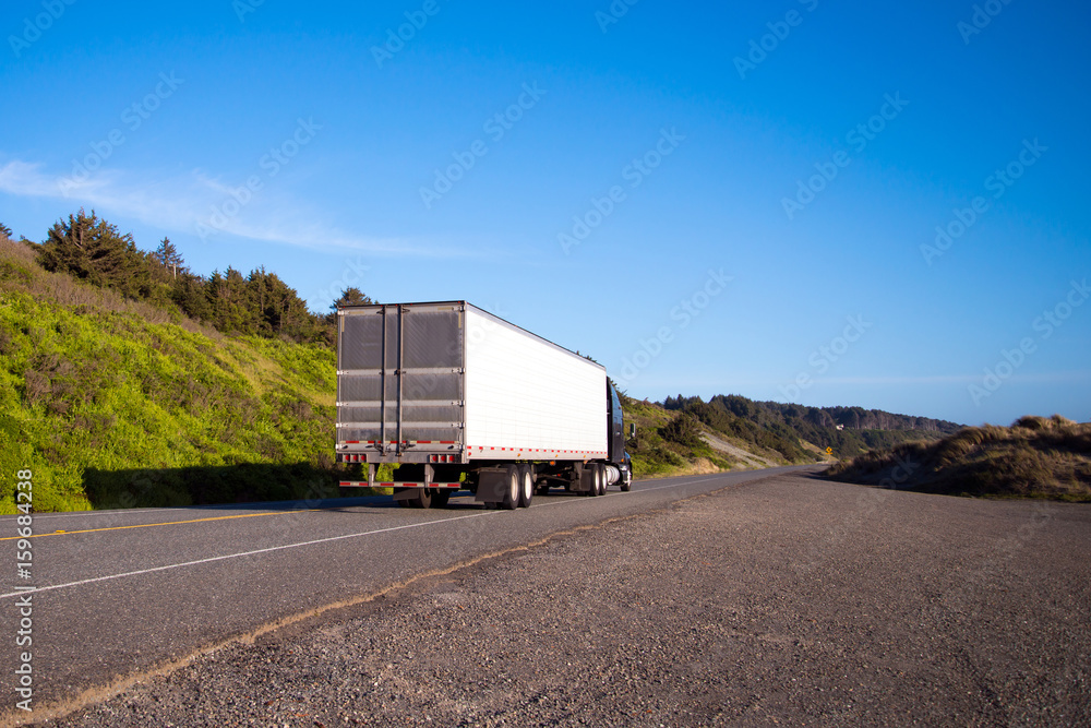 Black semi truck with trailer drive on straight road with green grass and bushes Black semi truck with trailer drive on straight road with green grass and bushes