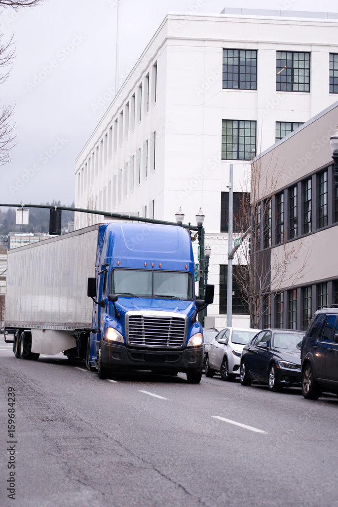 Blue big rig semi truck with trailer turn on the street of urban city Blue big rig semi truck with trailer turn on the street of urban city