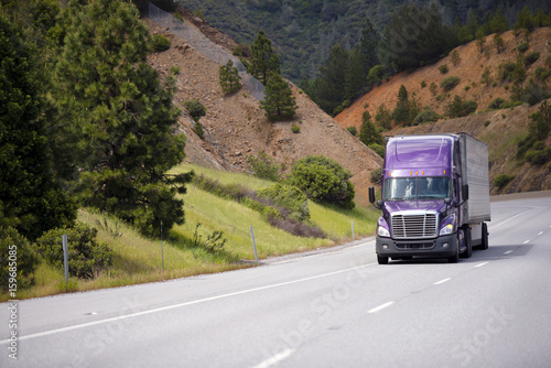 Lilac semi truck with aluminum trailer is moving along winding highway