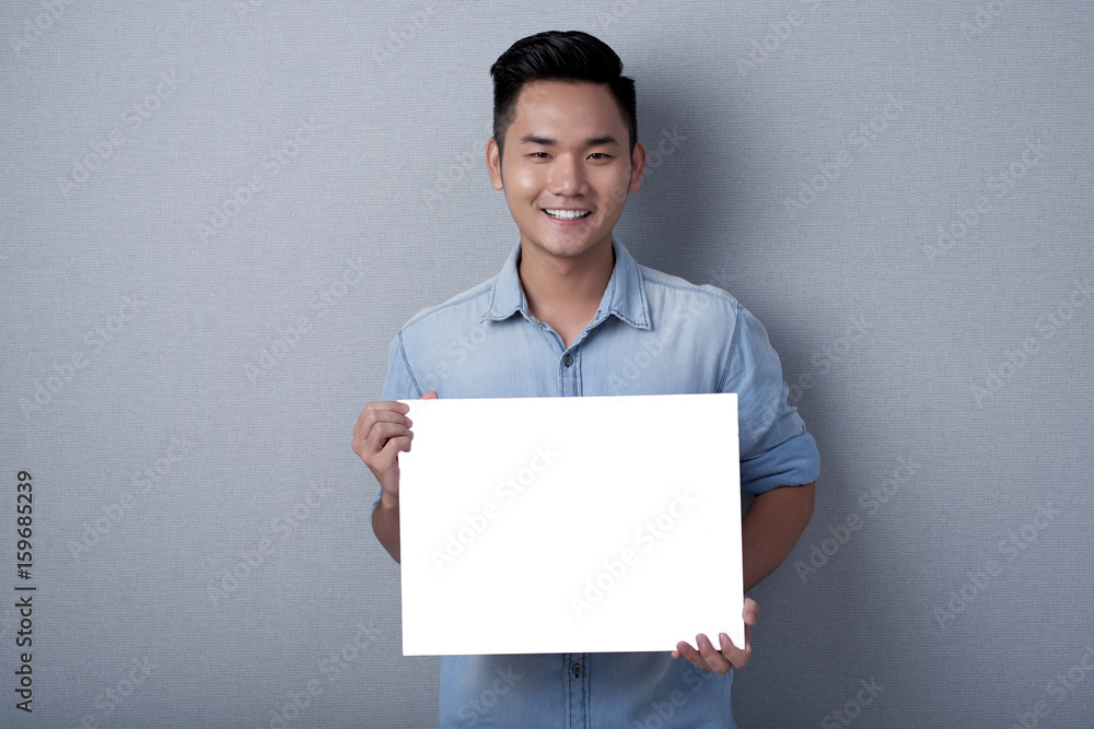 Joyful Asian man with stylish haircut posing for photography against gray background while holding blank sheet of paper in hands, studio shot Joyful Asian man with stylish haircut posing for photography against gray background while holding blank sheet of paper in hands, studio shot
