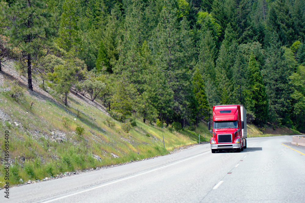 Modern red semi truck with trailer move cargo uphill on awesome green highway with forest trees on roadside hills Modern red semi truck with trailer move cargo uphill on awesome green highway with forest trees on roadside hills