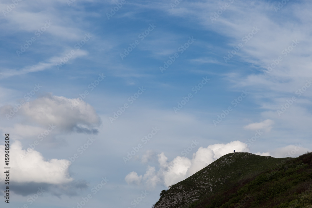 A small human figure on a cliff of a mountain beneath a huge blue sky with white clouds
