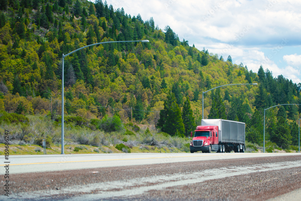 Red modern semi truck with low cab and soft tent trailer move on divided road with green trees on the hills Red modern semi truck with low cab and soft tent trailer move on divided road with green trees on the hills