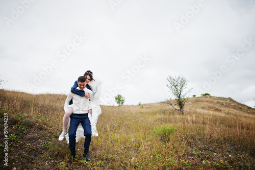 Happy bride riding her husband's back on their wedding day on the of hill with tall grass. © AS Photo Family