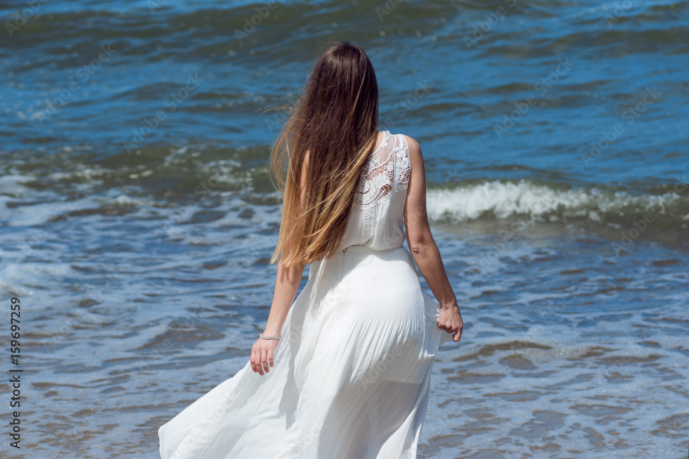 Young charming brunette woman on sea coast. Beautiful girl in a white summer dress. Runs towards the sea, back view