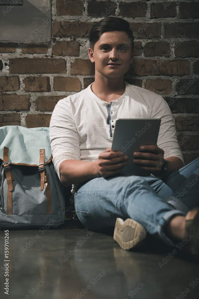 Man in casual clothes using digital tablet leaning to brick wall with backpack