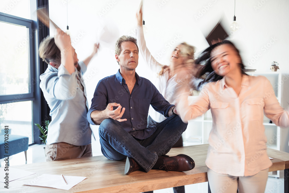 Relaxed businessman meditating in lotus position while coworkers celebrating success