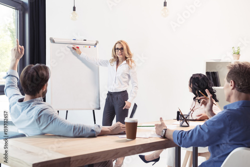 Blonde businesswoman in eyeglasses pointing at blank whiteboard and looking at colleagues