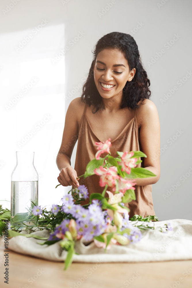 Cheerful beautiful african florist smiling making bouquet at workplace over white wall. Cheerful beautiful african florist smiling making bouquet at workplace over white wall.