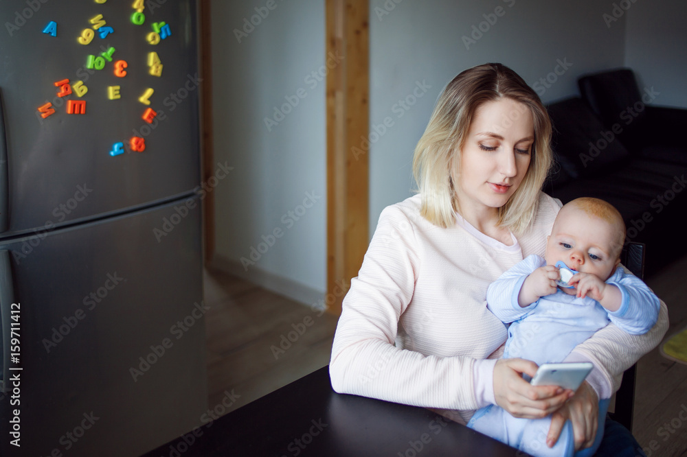 Young mother with baby sits and uses the phone. Internet browsing, indoor Young mother with baby sits and uses the phone. Internet browsing, indoor