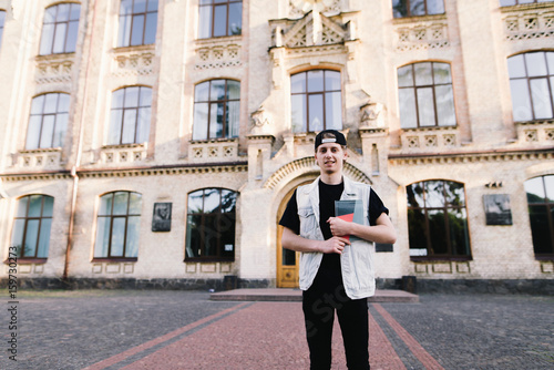 Portrait of a young man with a laptop in his hand standing at a college campus on the background of a building. A smiling student left the university. Student life.