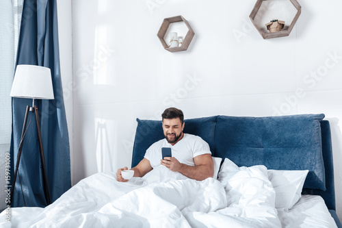 Young man using smartphone and having morning coffee in bed