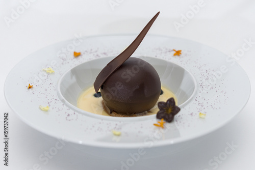 Luxurious dessert with chocolate globe and vanilla sauce  served on a biscuit  decorated with chocolate flower  flower petals and forest fruits  served in a white plate  light background  isolated