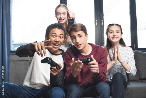 happy multicultural teenagers playing video games with joysticks at home, teenagers having fun concept