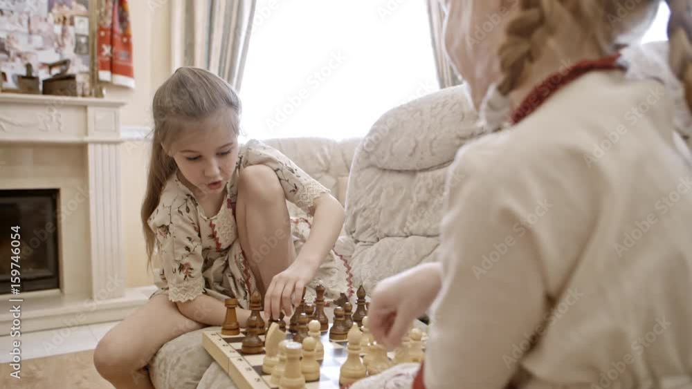 Cute girl in traditional Russian dress playing chess with sister on sofa at home  