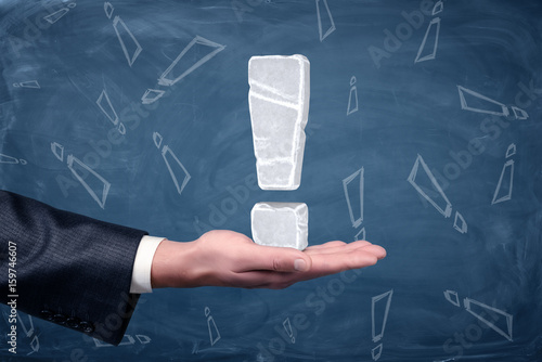 Φωτογραφία An exclamation mark made of white ice placed on businessman's open palm on chalkboard background