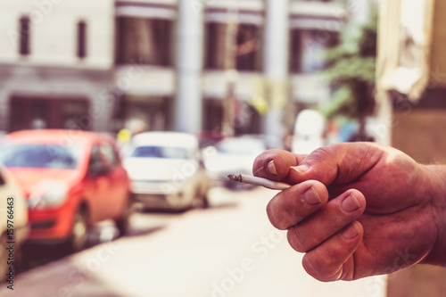 Man's hand holds a cigarette on a blurred city background, smoking in a public place © DedMityay