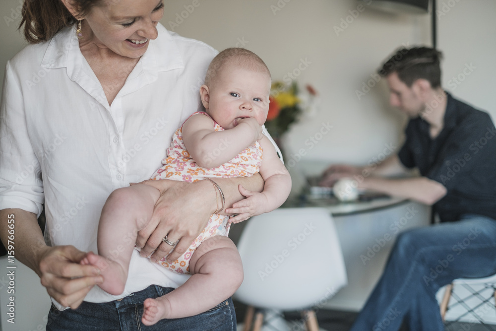Mother holding baby (2-5 months) and father working in background