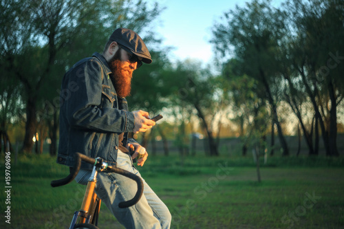 Bearded guy with mobile phone near fix bike. A brutal young man with a huge beard in sunglasses and a cap in the woods at sunset. Lifestyle.