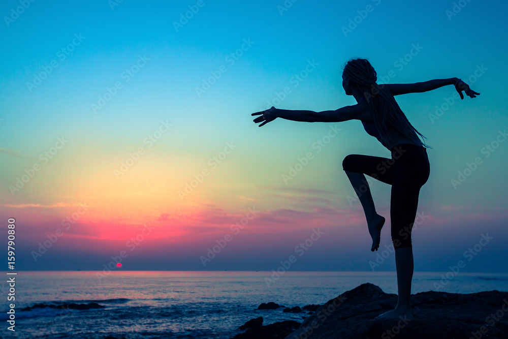 Silhouette of young flexible dancing woman on the sea coast during twilight.