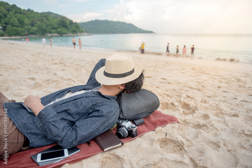 young Asian man lying on the beach and close his face by hat, vacation time and summer holiday concepts