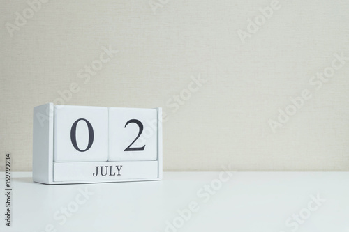 Closeup white wooden calendar with black 2 july word on blurred white wood desk and cream color wallpaper in room textured background with copy space , selective focus at the calendar