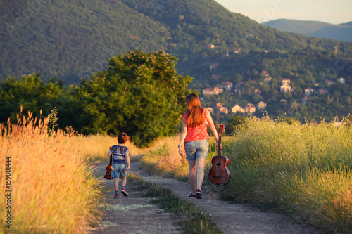 Woman with a little boy hold guitar and walking on rural road. Mom and her son leaving into the unknown in sunset