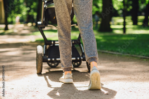 Young mother exercises in a park. Wears gray sport shoes, pushes a pram, close up, back view