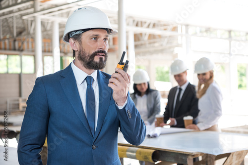 mature architect in suit talking on radio set during work at construction site