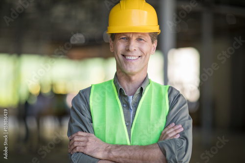 Professional mature architect in protective workwear standing with crossed arms and smiling at camera