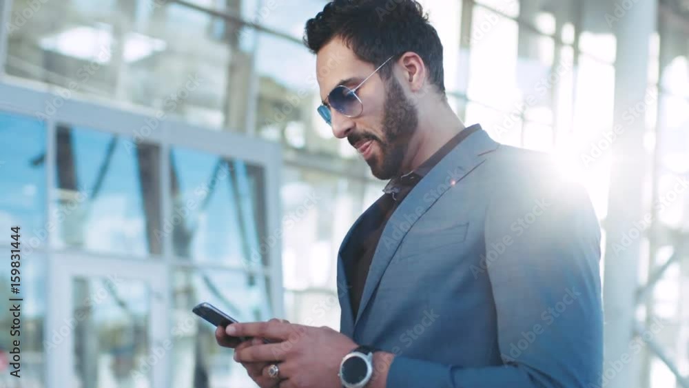 Handsome bearded Eastern businessman standing by the airport entrance, using his phone and smiles to received message, texts back. Stylish suit, sunglasses, elite watch. Successful lifestyle. Handsome bearded Eastern businessman standing by the airport entrance, using his phone and smiles to received message, texts back. Stylish suit, sunglasses, elite watch. Successful lifestyle.