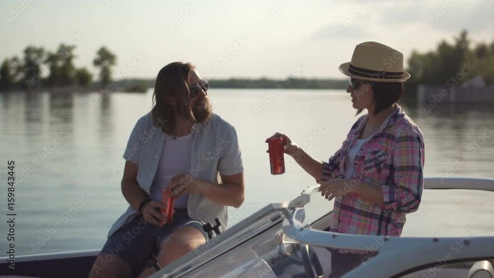 Young couple toasting each other with cans of beer as they relax in the evening on a lake in a motorboat while enjoying their summer vacation.