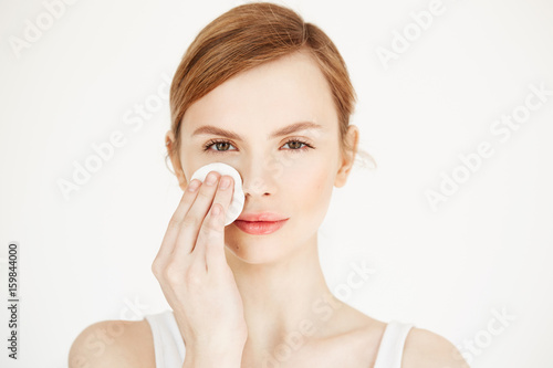 Beautiful natural blonde girl cleaning face with cotton sponge smiling looking at camera over white background. Cosmetology and spa.
