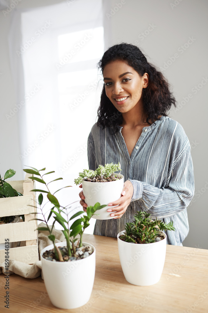 Beautiful african girl smiling looking at camera holding flower pot in hands over white wall. Beautiful african girl smiling looking at camera holding flower pot in hands over white wall.