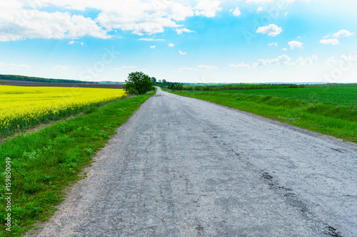 Old asphalt countryside road through green yellow fields and white clouds on blue sky in summer day. Horizontal background, scenic adventures travel concept. Lonely calm mood meditative nature.