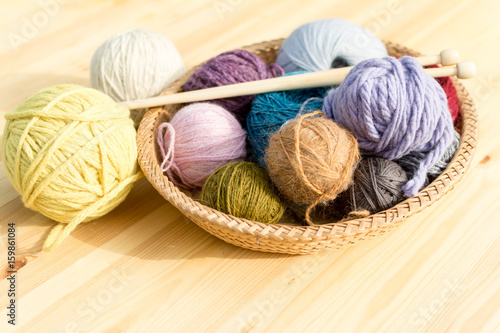 Set of colored yarn balls and needles on straw plate, wooden table as a background