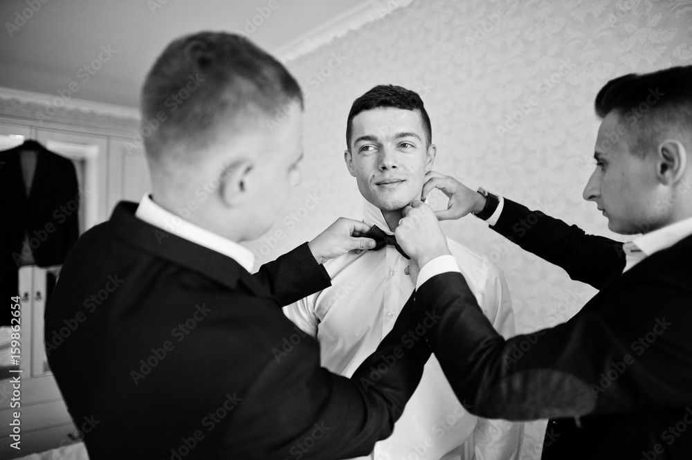 Groomsmen helping groom to dress up and get ready for his wedding in a room. Black and white photo. Groomsmen helping groom to dress up and get ready for his wedding in a room. Black and white photo.
